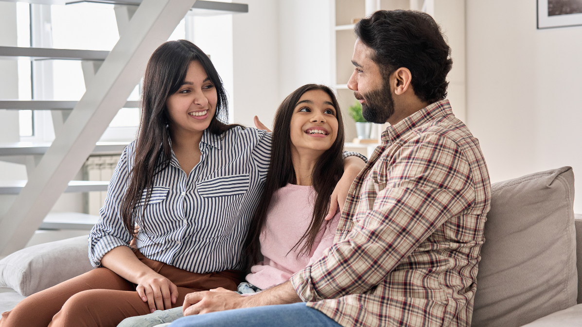 Two parents laughing and talking to their daughter on a sofa.