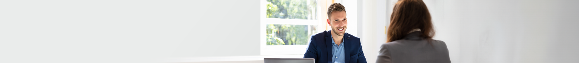 A white man smiling and interviewing someone at a desk. 