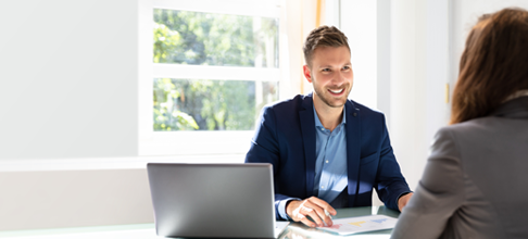 A white man smiling and interviewing someone at a desk.