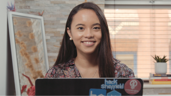 A woman stood in a bright room smiling over an open laptop which has stickers on the back.