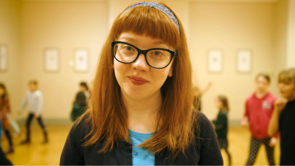 A woman standing and smiling in a room as children behind her take part in a drama class.