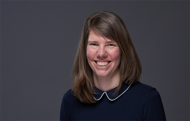 A woman with a dark blue blouse smiling widely in front of a dark background.