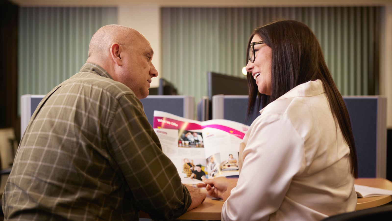 Two people are face to face, smiling and looking over a magazine which they are holding in front of them.