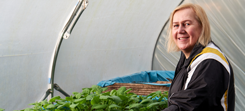 A lady looking into the camera and smiling, surrounded by plants in a greenhouse