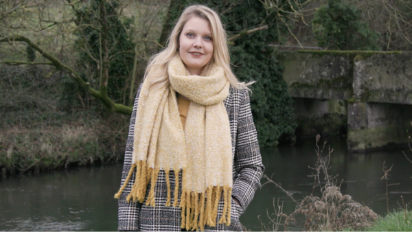 A woman with a coat and chunky scarf standing on a bank in front of a river.
