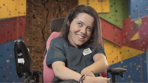 A woman in a wheelchair smiling in front of some colorful climbing walls.