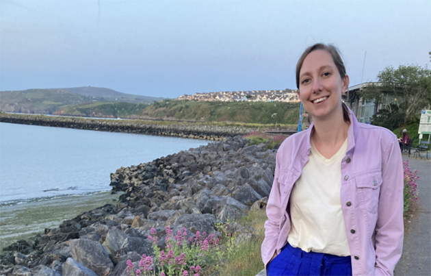 A woman with blue trousers and a pink jacket smiling on a coastal path.