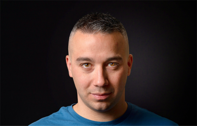 A man with a blue t-shirt is smiling in front of a dark background.